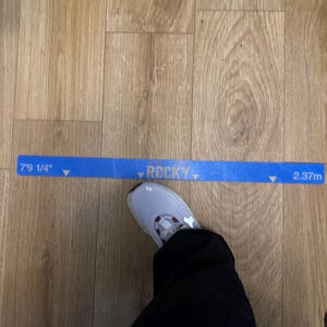 May include: A blue measuring tape with the word "ROCKY" and measurements of 7'9 1/4" and 2.37m. A person's foot in a gray and white athletic shoe is visible. The floor is made of wood.