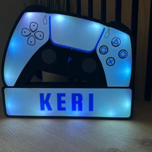 May include: An illuminated night light in the shape of a gaming controller, with blue LED lighting. The design features a black and white controller with the name "KERI" in blue. The controller has button details.