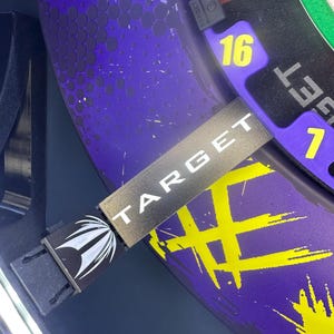 May include: Close-up of a dartboard with a purple and yellow design. The word "TARGET" is printed on a black and silver strap. The number 16 is visible in yellow. The dartboard has a black border.