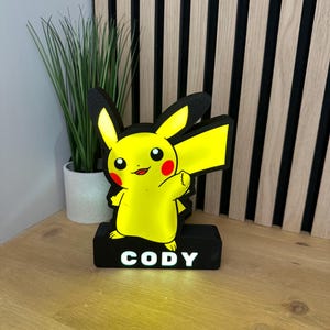 May include: A yellow Pikachu-shaped night light with black ears and a red cheek. The night light is mounted on a black base with the name "CODY" in white letters. A small green plant in a white pot is in the background.