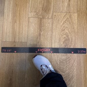 May include: A black measuring tape with the text "7'9 1/4" and "2.37m" in red. A white and burgundy athletic shoe is positioned near the tape. The tape also has the text "ASP" in red. The background is a wood-grain floor.