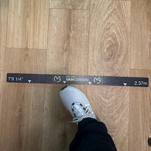 Op de afbeelding: Een zwarte meetlint met de tekst "MICHAEL VANGERWEN" en de afmetingen 7'9 1/4" en 2,37 m. De voet van een persoon in een witte en bordeauxrode sportschoen bevindt zich in de buurt van de tape op een houten vloer.