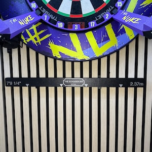 May include: A dartboard with a purple and yellow design, featuring the text "THE NUKE". The board is mounted on a wooden wall with a black bar that reads "THE STAGGER INN" and "7'9 1/4"" and "2.37m".