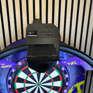 May include: A black and purple electronic dartboard with the text "THE NUKE" and "TARGET". The dartboard has a black, white, red, and green target area. A black electronic component is mounted above the dartboard.