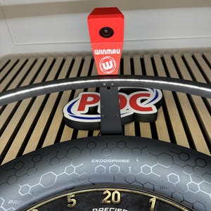 Könnte beinhalten: Eine schwarze Dartboard-Umrandung mit sechseckigem Muster und dem aufgedruckten Wort ENDORPHINE. Eine rote Winmau-Kameraeinheit ist oben montiert, positioniert über einem Dartboard mit goldenen Zahlen und dem Wort PRECISE auf der Oberfläche.