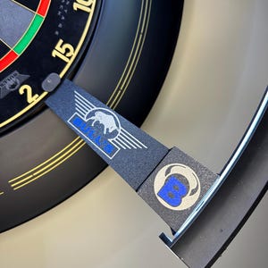 May include: A black dartboard surround ring featuring the Bulls Advantage Black logo in white and blue. The circular frame includes a rectangular extension piece with the brand name and a stylized bull graphic, designed to fit around a standard dartboard.