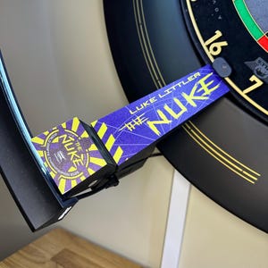 May include: A black dartboard with gold accents and a blue and yellow "The Nuke" dartboard surround. The surround features the text "Luke Littler" and "World Darts Champion 2023". The dartboard is mounted on a wall.