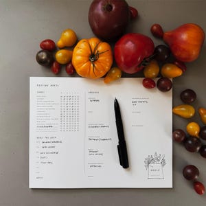 May include: A white paper planner titled Keeping House with columns for chores, groceries, and meals, featuring handwritten notes and a black ballpoint pen. The planner is surrounded by a variety of fresh, colorful heirloom tomatoes in shades of red, orange, and dark purple.