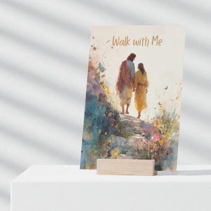 Op de afbeelding: Een aquarelprint met twee figuren die een stenen pad oplopen, met de woorden "Walk with Me" in gouden schrift. Het kunstwerk wordt gepresenteerd op een wit oppervlak met een houten standaard, met een sereen, spiritueel thema.