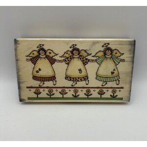 Puede incluir: Un sello de madera rectangular con una ilustración de tres ángeles tomados de la mano, cada uno con un vestido de diferente estampado. Debajo de los ángeles hay una fila horizontal de siete pequeños girasoles amarillos con tallos verdes.