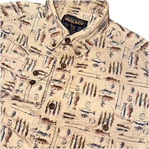 May include: Button-down shirt with a cream-colored background and a repeating pattern of fish, fishing rods, and flies. The shirt has a button-down collar, two chest pockets, and brown buttons. The brand label "Woolrich" is visible.
