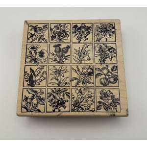 Pode incluir: Um carimbo de borracha quadrado de madeira com uma grade de dezasseis ilustrações individuais a tinta preta de vários temas botânicos, incluindo flores, bagas e folhas, dispostas em quatro filas e quatro colunas num bloco de madeira clara.