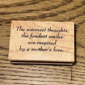 以下が含まれることがあります： 「The warmest thoughts, the fondest smiles are inspired by a mother's love」と書かれた長方形の木製ブロック。テキストは黒色のエレガントなフォントです。木材は明るい茶色で、木目が見えます。