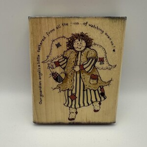 May include: A rectangular wooden rubber stamp featuring a line art illustration of an angel with wings, wearing a striped dress and a textured coat with patches. The text reads: Our guardian angel's a little tattered from all the fuss... of watching over us!