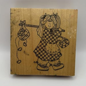 Puede incluir: Un sello de goma cuadrado de madera con una ilustración de líneas negras de una persona con coletas que lleva un vestido a cuadros y un palo sobre el hombro con un hatillo atado, del cual caen varios corazones pequeños.