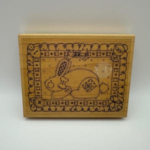 May include: A rectangular wooden rubber stamp featuring a black line art illustration of a rabbit. The rabbit is surrounded by a decorative border composed of buttons, ribbons, and scalloped edges. The design includes a bow and a patch detail.