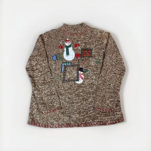 May include: Brown knit sweater with a mock turtleneck and red stitching. The sweater features appliqued snowmen, hearts, and a plaid square. The design includes a blue and green Christmas tree and a small blue ornament.