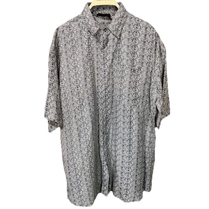 Op de afbeelding: Kort mouwshirt met een herhalend cirkelvormig patroon in grijstinten en zwart. Het shirt heeft een klassieke kraag en een borstzak. Het kledingstuk hangt aan een houten kleerhanger tegen een witte achtergrond.