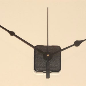 May include: Black clock hands with a square black base. The hands are made of metal and have a pointed tip.