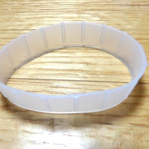 May include: A translucent, ring-shaped plastic object with vertical indentations. The object is positioned on a wooden surface, and appears to be a component or part of a larger item. The ring is approximately 3 inches in diameter.