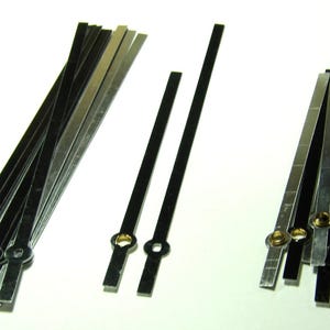 May include: A collection of long, thin, straight clock hands in black and silver metallic finishes. The hands feature circular mounting holes at one end and vary in length, with some arranged in a loose fan shape and others standing separately.
