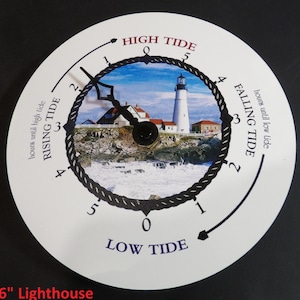 May include: A white circular tide clock with a black rope design around the edge. The clock face features a lighthouse and ocean scene. The clock has markings for high tide, low tide, rising tide, and falling tide. The text "6" Lighthouse" is printed below the clock.