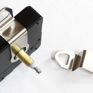 May include: A black clock movement with a silver metal bracket and a separate silver metal bracket with a hole. The clock movement has a gold-colored shaft with a white tip.