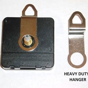 May include: A black clock movement with a gold-colored hanger. The movement has a small hole in the center for the clock hand. The hanger is labeled "Heavy Duty Hanger".