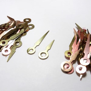 Puede incluir: Un montón de manecillas de reloj doradas con una punta puntiaguda y un agujero circular en la base. Hay dos manecillas de reloj individuales en el centro de la imagen.
