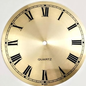 May include: A circular gold-toned clock dial featuring black Roman numerals from I to XII. The center has a small circular hole for clock hands. The word QUARTZ is printed in black above the center, and the text MCCI is printed at the bottom edge.