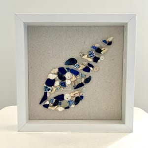 May include: A framed artwork featuring a stylized leaf design composed of various blue, white, and beige ceramic pieces and shells. The artwork is set against a neutral linen background within a white wooden frame, creating a coastal aesthetic.