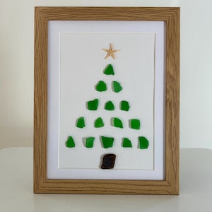 May include: Framed artwork depicting a Christmas tree crafted from green sea glass. A starfish adorns the top, with a brown piece of glass as the trunk. The design is set on a white background, encased in a wooden frame.