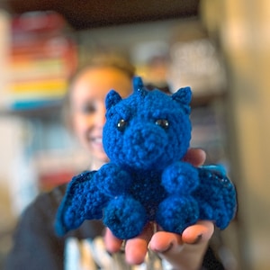 May include: A blue crocheted dragon toy held in a person's hand. The dragon has a small, rounded body with a long, pointed snout and large, black eyes.