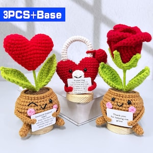 Puede incluir: Tres decoraciones de crochet con forma de corazón y flor. El conjunto incluye un corazón rojo, un candado en forma de corazón y una rosa roja. Cada uno se encuentra en una maceta marrón con una cara sonriente y sostiene un pequeño letrero con texto.