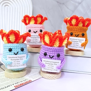 Puede incluir: Cuatro peluches de ganchillo con forma de fuego con una cara sonriente y sosteniendo un cartel que dice "Emotional Support Dumpster Fire It's fine, I'm fine. Everything is fine!" Los peluches son de color rosa, naranja, azul claro y morado.