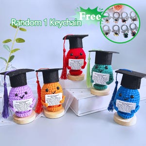 Puede incluir: Una colección de figuras de ganchillo coloridas, cada una con un birrete negro y una borla. Las figuras son de color púrpura, naranja, rojo, verde y azul, y están montadas sobre pequeñas bases de madera. Cada figura tiene una cara sonriente y un pequeño letrero.