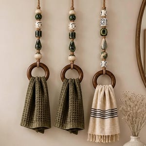 Peut inclure: Trois porte-serviettes muraux composés d'anneaux en bois foncé suspendus par des cordes de jute ornées de perles en céramique décoratives dans des tons vert, crème et noir. Deux supports présentent des serviettes gaufrées vert olive, tandis que le troisième porte une serviette crème à rayures noires et franges.