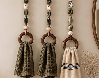 May include: Three wall-mounted towel holders featuring dark wooden rings suspended by jute ropes adorned with decorative ceramic beads in shades of green, cream, and black. Two holders display olive green waffle-weave towels, while the third holds a cream towel with black stripes and fringe.