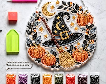 May include: A circular diamond painting kit featuring a witch hat, broom, and pumpkins surrounded by foliage. Includes a pink stylus, a green tray, a wax square, a metal hanging chain, and eight small bags of colorful resin beads for assembly.