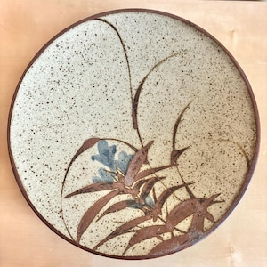 Pode incluir: Prato redondo de cerâmica com superfície bege salpicada e borda castanha escura. O prato apresenta um desenho floral com folhas castanhas e flores azuis. O desenho é centrado, com linhas castanhas.