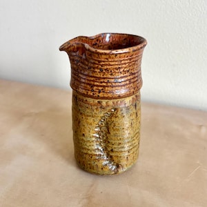 以下が含まれることがあります： 注ぎ口のある円筒形の陶器製クリーマー。上部は茶色の斑点模様の釉薬で覆われ、下部はテクスチャーのあるマスタードイエローで、容器の外側表面には縦方向のくぼんだ模様が施されています。