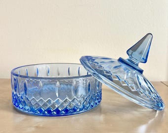 May include: A blue glass trinket dish with a lid. The dish is round with a diamond pattern on the sides and a pointed finial on the lid. The lid is slightly ajar, revealing the interior of the dish.