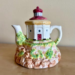 May include: A ceramic teapot shaped like a lighthouse. The teapot features a white tower with a red roof, a red door, and small blue windows. The base is a rocky, textured design with green and yellow accents.