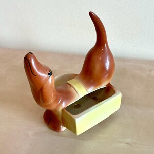 Könnte beinhalten: Keramik-Dackel-Figur in Braun- und Gelbtönen. Der Hund hat Kopf und Schwanz erhoben, mit einem rechteckigen Pflanzgefäß. Die Figur hat schwarze Akzente und eine glänzende Oberfläche. Der Pflanzkasten ist ca. 10 cm lang.