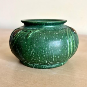 May include: A round, squat ceramic bowl with a deep forest green glaze featuring a mottled, textured finish. The exterior surface displays raised, organic relief patterns resembling vines or leaves, with subtle brown accents on the raised details.