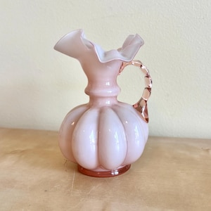 May include: A pink glass pitcher with a ruffled rim and a bulbous body. The handle is a lighter shade of pink and has a twisted design. The pitcher sits on a round base and is a decorative item.