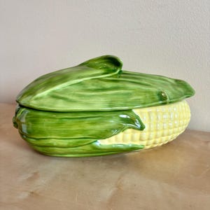 Puede incluir: Un plato de servicio de cerámica con forma de mazorca de maíz y tapa extraíble. La tapa presenta un acabado verde brillante que imita las hojas de maíz, mientras que la base muestra un patrón amarillo texturizado de granos.