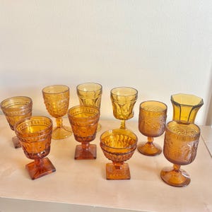 Może przedstawiać: Kolekcja szklanych naczyń w stylu vintage w kolorze bursztynowym, w tym kielichy i kieliszki. Szklanki mają różne wzory, takie jak teksturowane wzory i motywy kwiatowe. Szkło jest ułożone na jasnej powierzchni.