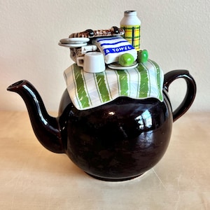 Puede incluir: Una tetera de cerámica marrón oscuro con un diseño caprichoso. La tapa presenta objetos en miniatura como una toalla con el texto "A TOWEL", una botella y una tela a rayas. La tetera tiene un pico y un asa curvados, con un acabado brillante.