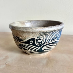 May include: A ceramic bowl featuring a speckled beige upper rim and a dark blue lower section decorated with white etched line patterns. The bowl has a rounded shape with a smooth, curved edge and a visible contrast between the textured clay.